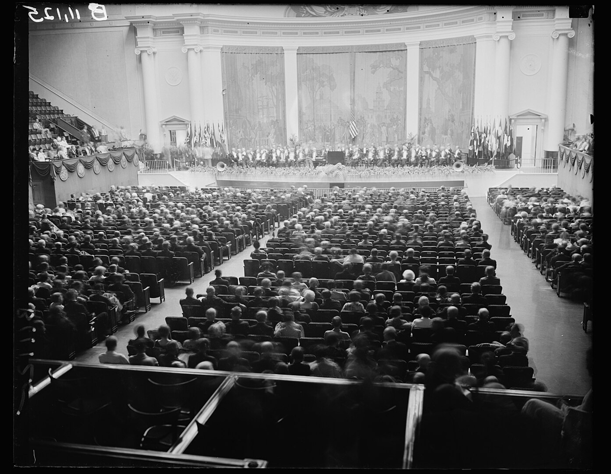 Bestuhlter großer Saal mit Hunderten von Menschen; vorne eine Bühne mit Rednern und Fahnen.