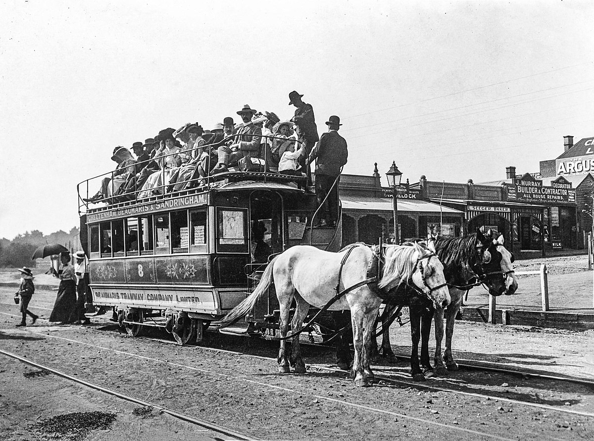 Pferdegezogene Straßenbahn Ein Wagen, auf dem oben Menschen im Freien sitzen, wird von zwei Pferden gezogen.