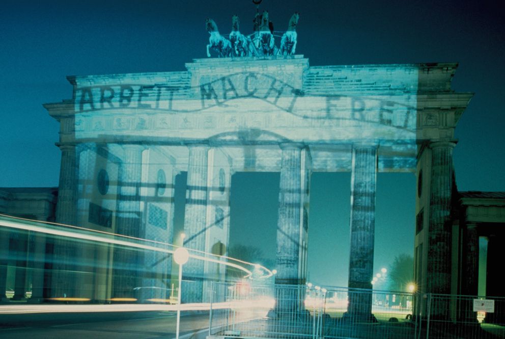 Fotografie eines großen Monuments mit Säuen. Es wird mit einem Bild bestrahlt auf dem steht: "Arbeit macht frei". Dahinter eine Straße.