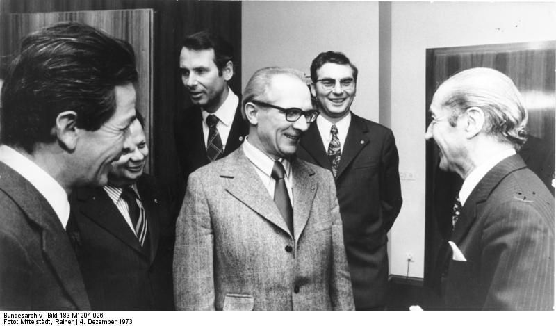 Schwarz-Weiß Fotografie einer Gruppe anzugtragender Männer. Sie lächeln sich gegenseitig an.