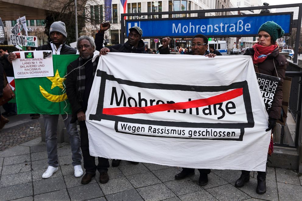 Fotografie einer gruppe Menschen vor dem Ubahn Ausgang der Haltestelle "Mohrenstraße". Sie halten ein Banner auf dem das besagte Haltestellenschild durchgestrichen ist. Daneben die Mauretanische Nationalflagge, mit der Aussage: Nummer 1 Land in Sklaverei und Rassismus gegen Schwarze.