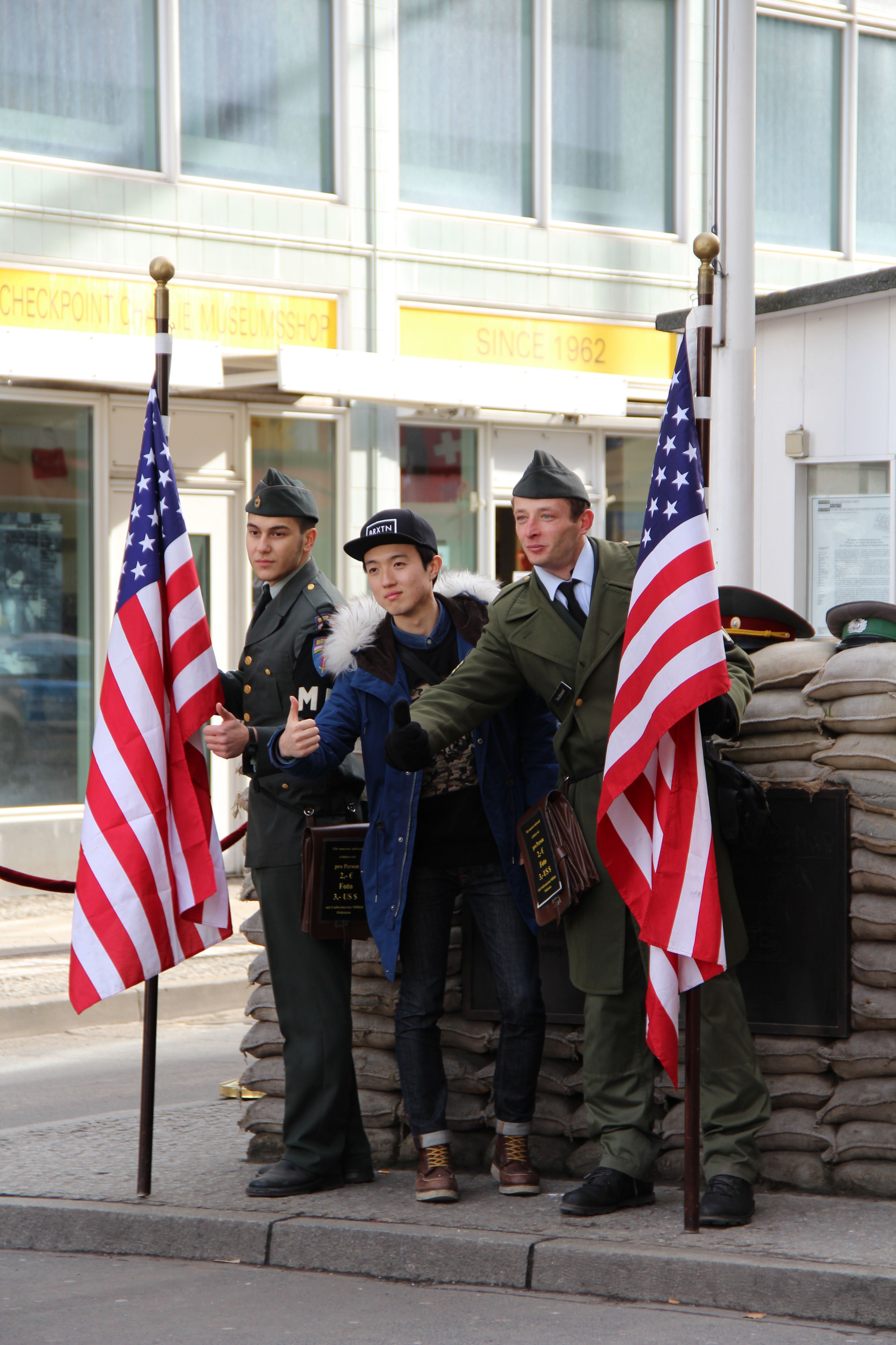 Foto von drei Männern vor einer Wand aus Sandsäcken. Zwei davon in Uniformen und USA Flaggen in der Hand, einer modern gekleidet.