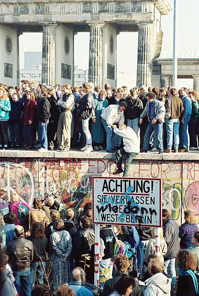 9. November 1989: Menschen auf der Berliner Mauer vor dem Brandenburger Tor. Die Aufschrift auf dem Schild: „Achtung! Sie verlassen jetzt West-Berlin“ wurde übersprüht mit der Frage „Wie denn?“. Fotograf: Sue Ream (San Francisco, California), Quelle ([https://creativecommons.org/licenses/by/3.0/deed.en CC BY 3.0]): [http://commons.wikimedia.org/wiki/File:BerlinWall-BrandenburgGate.jpg Wikimedia Commons].