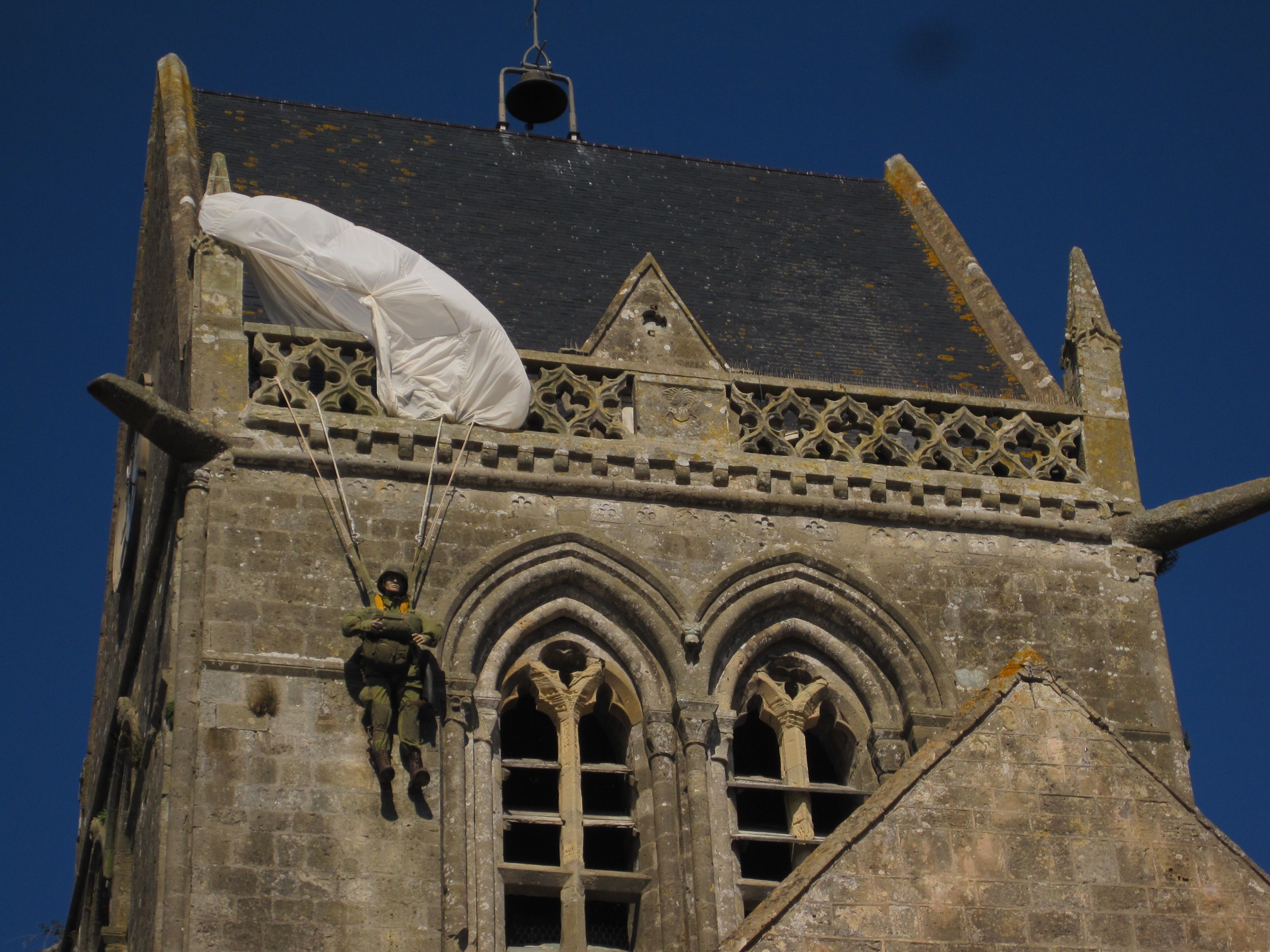 Eine andere Form der öffentlichen Geschichtsrepräsentation stellt diese Fallschirmspringer-Puppe am Turm der Kirche von Sainte-Mère-Église in der Normandie in Frankreich dar. Sie soll an einen amerikanischen Soldaten erinnern, der bei der alliierten Invasion 1944 versehentlich an diesem Kirchturm hängenblieb. Dieser eher spielerische Zugang zeigt einen relativ ungezwungenen Umgang mit Geschichte, wirkt die Puppe doch fast wie eine Werbemaßnahme für das gegenüber der Kirche stehende Airborne Museum. 
Foto: Irmgard Zündorf, Sainte-Mère-Église 2015, Lizenz: [https://creativecommons.org/licenses/by-nc/3.0/de/ CC BY-NC 3.0 DE]