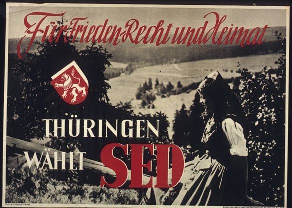 The SED in Thuringia, East Germany, canvasses votes with a photographic image that could have just as well been used on movie posters for a West German Heimatfilm. A young woman in what appears to be traditional costume sits on a wooden fence before a sun-drenched valley free of modern influences. The poster reveals that older (bourgeois) notions of Heimat were still commonplace in the GDR. Only with the advent of a “socialist Heimat” in the 1950s did an alternative program emerge.
<br />
SED election poster: “For peace, justice and homeland,” SED state executive committee of Thuringia, September/October 1946 ca. 42 x 58 cm. Source: Bundesarchiv Plak 100-015-038 courtesy of the German Federal Archives