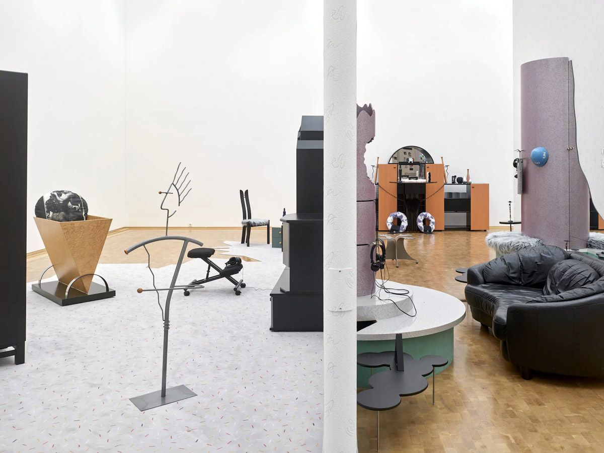 Installationsansicht des Projekts „2000“ der Künstlerin Henrike Naumann (https://henrikenaumann.com/work/2000/): ein deutsch-deutscher Pavillon der Postmoderne gefüllt mit Objekten und Möbelstücken. Ausstellung im Museum Abteiberg, Mönchengladbach, 11.03.2018-10.06.2018. Foto: Achim Kukulies © Henrike Naumann <br />Henrike Naumann reflektiert gesellschaftspolitische Probleme auf der Ebene von Design und Interieur und erkundet das Reibungsverhältnis entgegengesetzter politischer Meinungen im Umgang mit Geschmack und persönlicher Alltagsästhetik. Sie hat das Jahr „2000“ als Ausgangspunkt genommen für eine Betrachtung von Ästhetik und Gesellschaft seit der deutschen Einheit in Ost- und Westdeutschland: Gibt es einen Zusammenhang von Design und Politik? Welche gesellschaftlichen Auswirkungen hatte der postmoderne Bauboom ab 1990 für das Leben der Menschen in der ehemaligen DDR? Kann man sich durch Möbel radikalisieren?