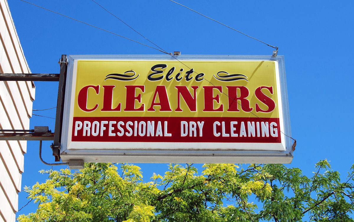Fotografie eines gelben Leuchtkastenschilds. Darauf in roter und schwarzer Schrift: Elite Cleaners. Im Hintergrund blauer Himmel und ein grüner Baum.