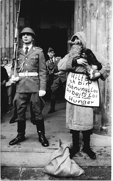 Schwarz-weiß Foto zweier Menschen.n Links: ein Man in Uniform mit einem Gewehr. Rechts: Mann mit langem Bart und einem umgehängte Schild mit der Aufschrift: Hilfe ich bin Wohnungslos Arbeitslos und habe Hunger.