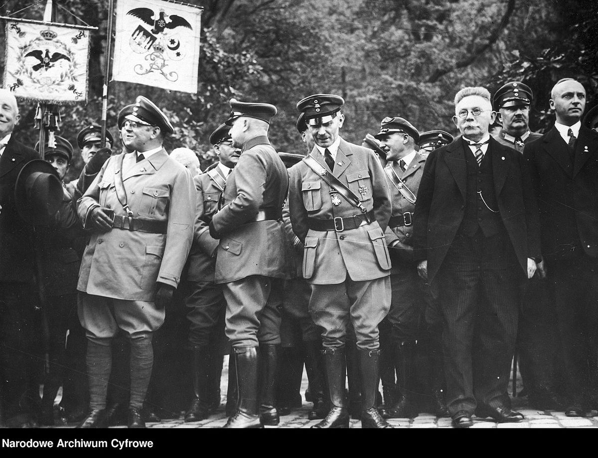 Schwarz-Weiß-Fotografie einer Gruppe von weißen Männern. Teile in Uniform teile in schwarzen Anzügen. Es werden zwei Standarten mit einerm Wappen gehalten. Auf dem Wappen unter anderem ein Vogel.