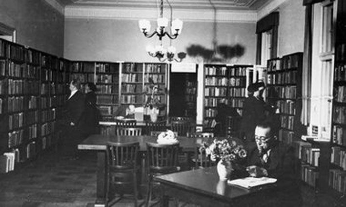 Schwarz weiß Foto eines Raums voller Bücherregale vor denen Menschen stehen. In der Mitte drei Tische. An einem sitzt ein lesender Mann.
